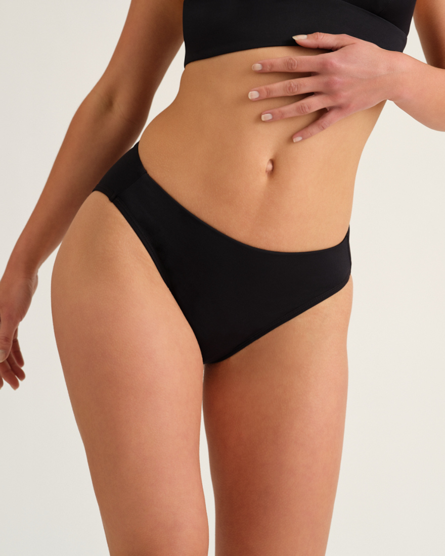 Modell trägt den ooia Beach Slip in Schwarz - Periodenbikini-Slip im klassischen Design