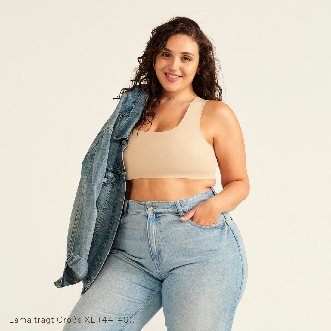 Modell trägt ooia All-You-Need Bra Top in XL in Beige – bügelloses, nahtloses Bustier mit breiten Trägern, als Top zu Jeans und Jacke.