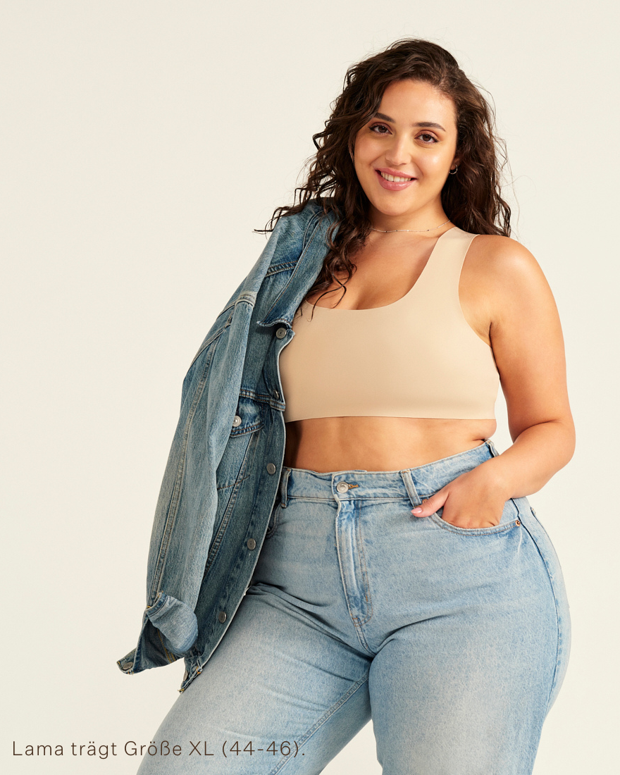 Modell trägt ooia All-You-Need Bra Top in XL in Beige – bügelloses, nahtloses Bustier mit breiten Trägern, als Top zu Jeans und Jacke.
