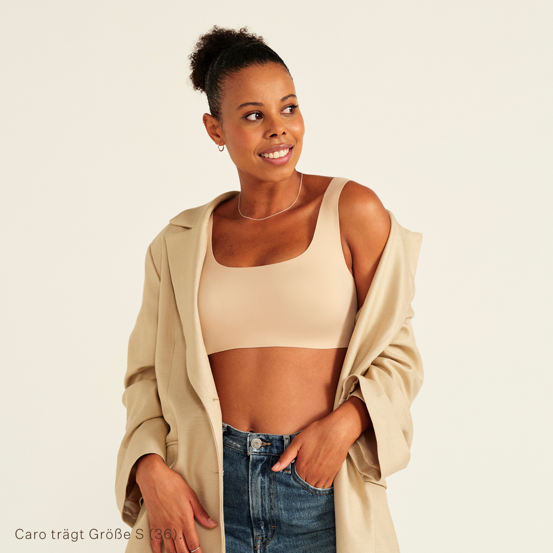 Modell trägt ooia All-You-Need Bra Top in XL in Beige – bügelloses, nahtloses Bustier mit breiten Trägern, als Top zu Jeans und Jacke.