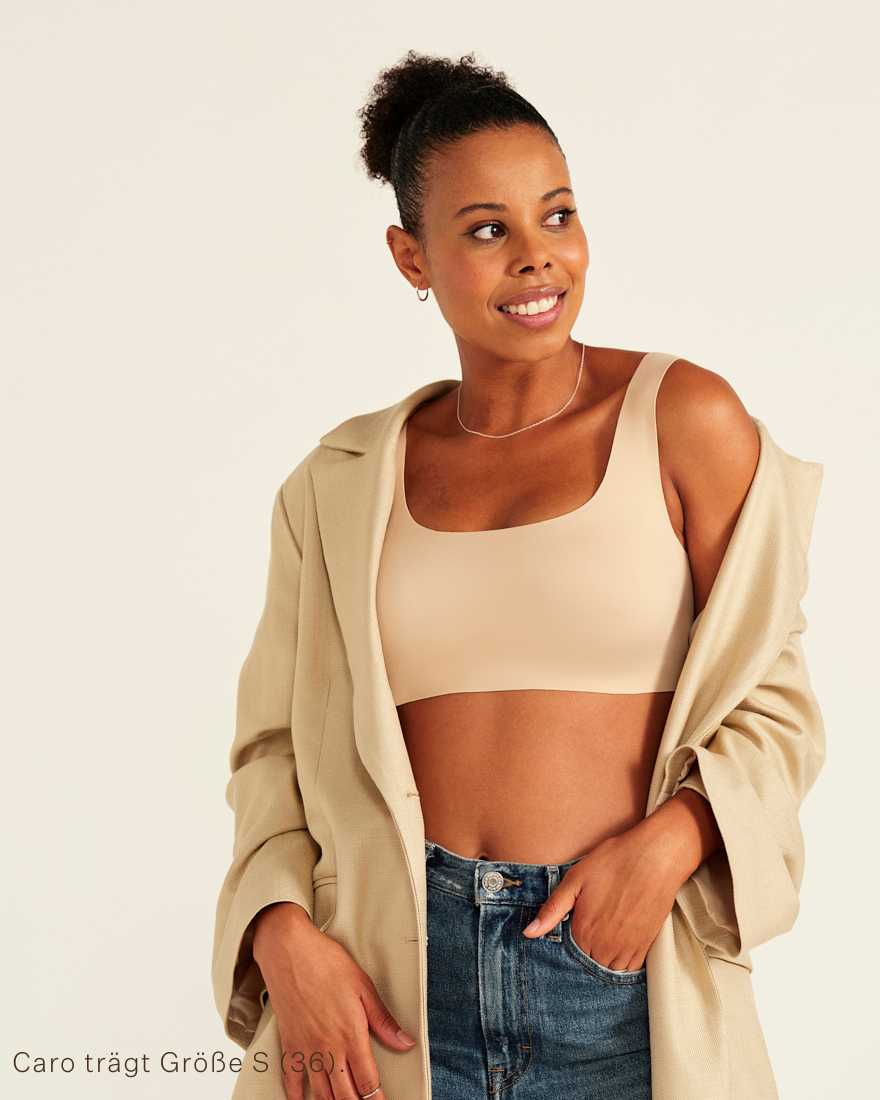 Modell trägt ooia All-You-Need Bra Top in XL in Beige – bügelloses, nahtloses Bustier mit breiten Trägern, als Top zu Jeans und Jacke.