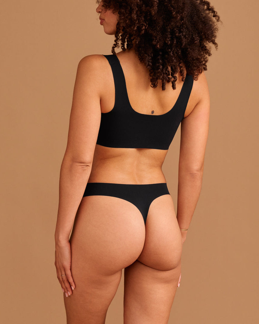 Modell trägt ooia Periodenunterwäsche String Seamless in Schwarz - nahtloser String mit leichter Saugstärke. Rückansicht.