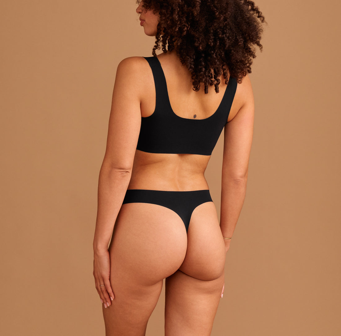 Modell trägt ooia Periodenunterwäsche String Seamless in Schwarz - nahtloser String mit leichter Saugstärke. Rückansicht.