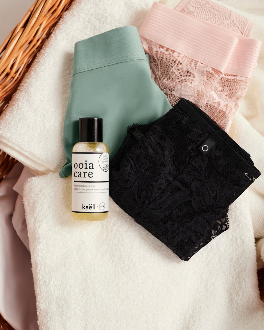 ooia Care Waschmittelkonzentrat für schonende Reinigung in kleiner Flasche, liegt auf einen Wäschekorb mit weißen Handtüchern und ooia Periodenunterwäsche in Sschwarz, Sage und Powder Rose