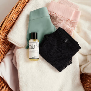 ooia Care Waschmittelkonzentrat für schonende Reinigung in kleiner Flasche, liegt auf einen Wäschekorb mit weißen Handtüchern und ooia Periodenunterwäsche in Sschwarz, Sage und Powder Rose