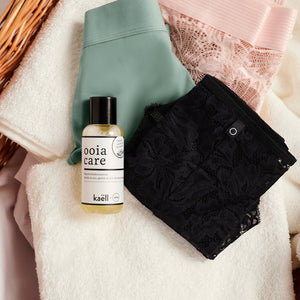 ooia Care Waschmittelkonzentrat für schonende Reinigung in kleiner Flasche, liegt auf einen Wäschekorb mit weißen Handtüchern und ooia Periodenunterwäsche in Sschwarz, Sage und Powder Rose