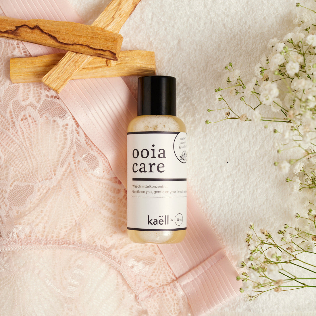 ooia Care Waschmittelkonzentrat für schonende Reinigung in kleiner Flasche, mit ooia Periodenunterwäsche, Zederholz und Blumen im Hintergrund.
