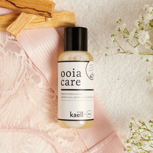 ooia Care Waschmittelkonzentrat für schonende Reinigung in kleiner Flasche, mit ooia Periodenunterwäsche, Zederholz und Blumen im Hintergrund.