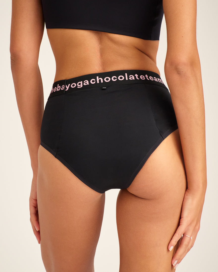 Modell trägt ooia Slip Hugs in Schwarz – bequemer Slip mit extra stützendem Bund mit Aufdruck und integrierten Taschen für das ooia Wärmepad. Rückansicht.