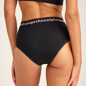Modell trägt ooia Slip Hugs in Schwarz – bequemer Slip mit extra stützendem Bund mit Aufdruck und integrierten Taschen für das ooia Wärmepad. Rückansicht.