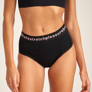Modell trägt ooia Slip Hugs in Schwarz – bequemer Slip mit extra stützendem Bund mit Aufdruck und integrierten Taschen für das ooia Wärmepad.