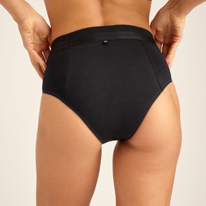Modell trägt ooia Slip Hugs Extra Strong in Schwarz – bequemer Slip mit extra stützendem Bund, starker Sauglstärke und integrierten Taschen für das ooia Wärmepad. Rückansicht.