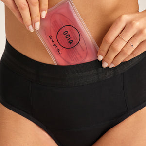 Modell trägt ooia Slip Hugs Extra Strong in Schwarz – bequemer Slip mit extra stützendem Bund, starker Sauglstärke und integrierten Taschen für das ooia Wärmepad. Model tut das Wärmepad in die vordere Tasche.