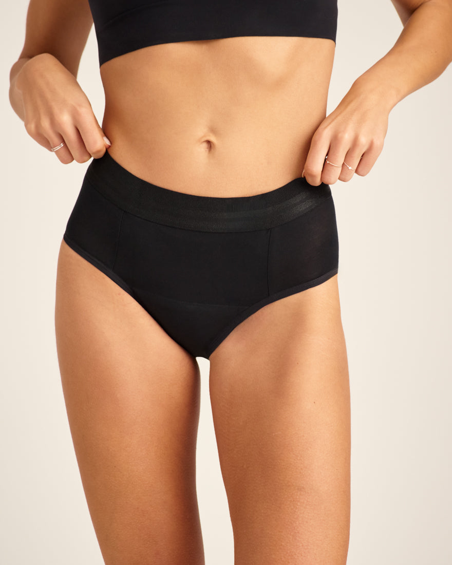 Modell trägt ooia Slip Hugs Extra Strong in Schwarz – bequemer Slip mit extra stützendem Bund, starker Sauglstärke und integrierten Taschen für das ooia Wärmepad.