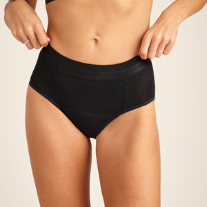 Modell trägt ooia Slip Hugs Extra Strong in Schwarz – bequemer Slip mit extra stützendem Bund, starker Sauglstärke und integrierten Taschen für das ooia Wärmepad.