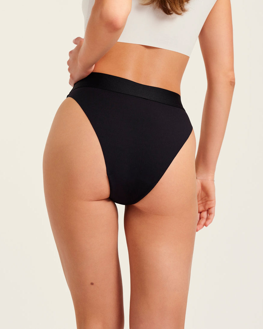 Modell trägt ooia Slip Seamless Cheeky in Schwarz – nahtlose Periodenunterwäsche mit hohem Beinausschnitt. Rückansicht.