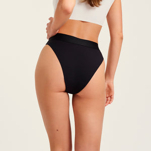 Modell trägt ooia Slip Seamless Cheeky in Schwarz – nahtlose Periodenunterwäsche mit hohem Beinausschnitt. Rückansicht.