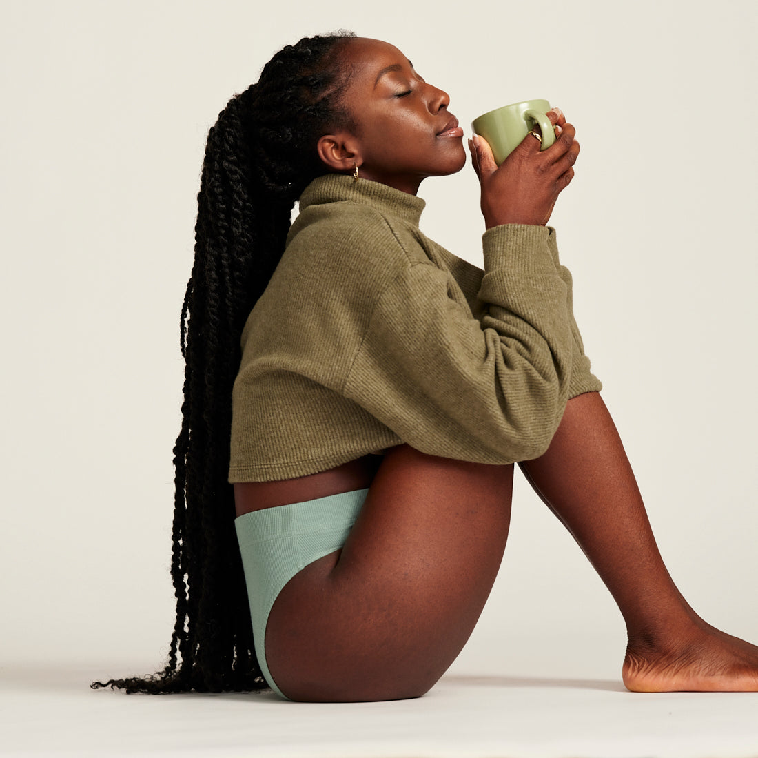 Model in grüner nahtloser Periodenunterwäsche und grünem Pullover sitzt auf dem Boden und trinkt Tee
