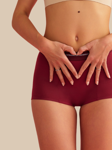 Ausschnitt eines Models, das eine rote Perioden-Shorts von ooia trägt und ein Herz mit ihren Händen auf ihrem Unterbauch formt.