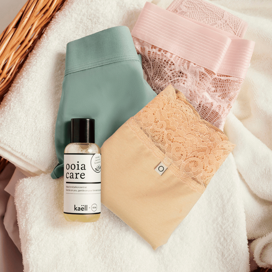 Drei verschiedenfarbige Periodenslips liegen mit ooia Waschmittelkonzentrat in einem Wäschekorb.