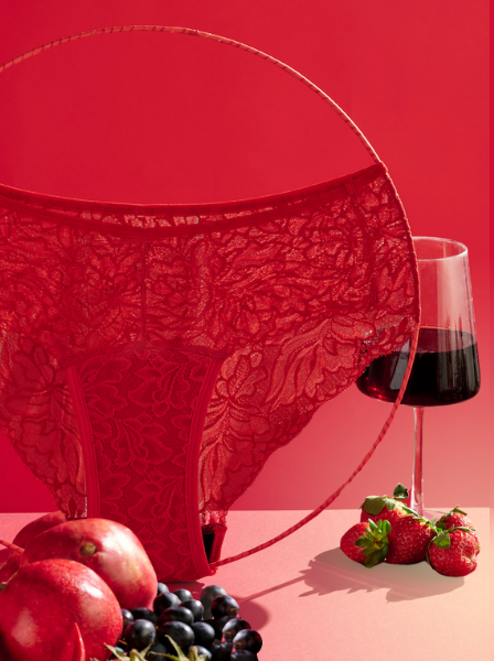 Der Periodenslip Hipster Allover Lace in Rot steht auf einem Tisch mit Rotwein, Erdbeeren, Äpfeln und weiterem roten Obst und Gemüse.