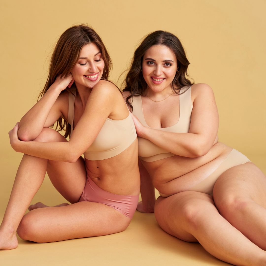 Zwei Models sitzen zusammen auf dem Boden und tragen nahtlose Periodenunterwäsche in rosa und beige
