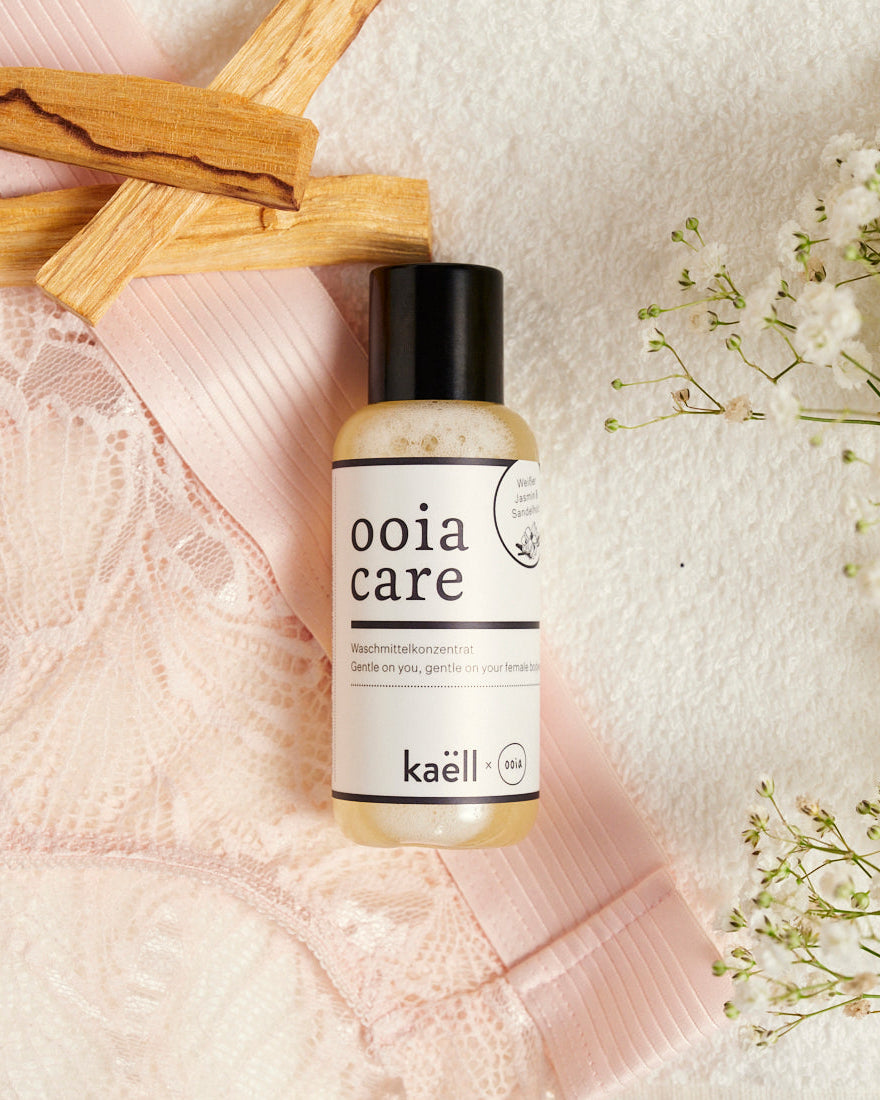 ooia Care Waschmittelkonzentrat für schonende Reinigung in kleiner Flasche, mit ooia Periodenunterwäsche, Zederholz und Blumen im Hintergrund.