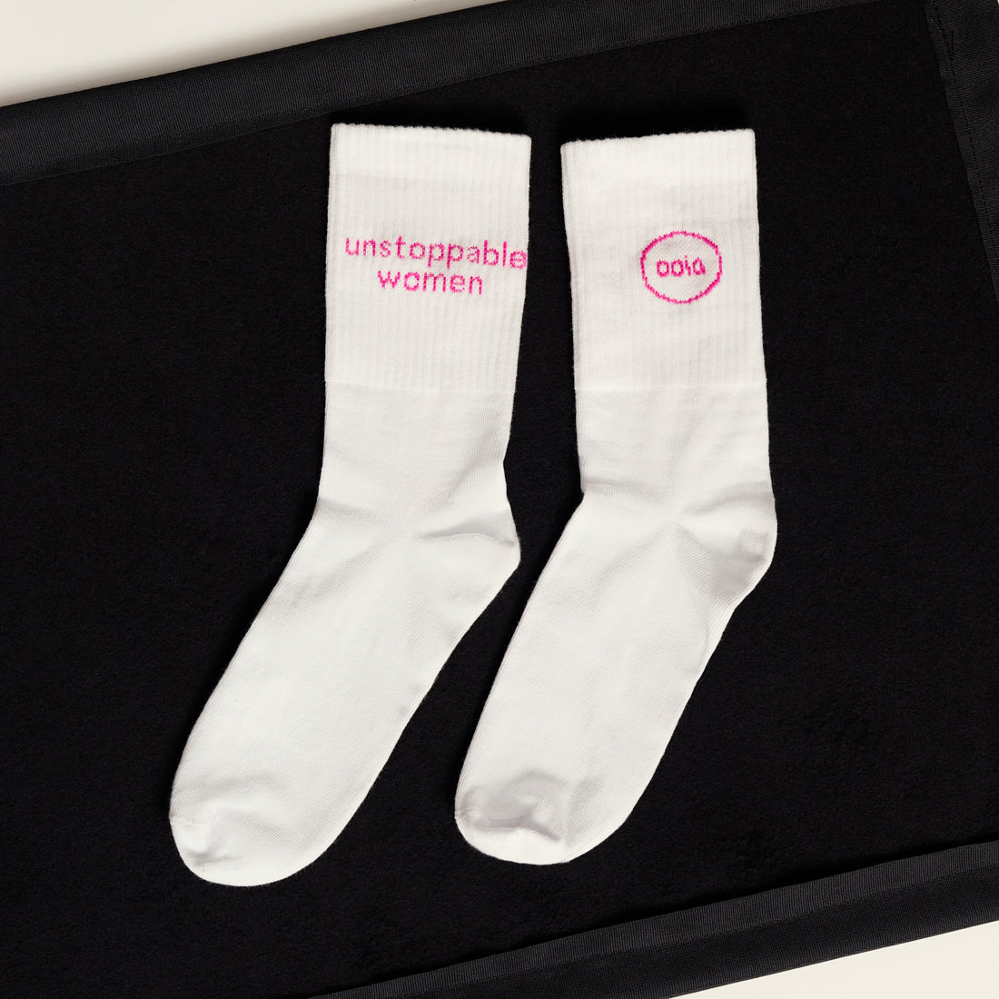 weiße ooia Socken mit dem pinken Aufdruck: Unstoppable Woman, liegen auf einer dunklen Fläche 