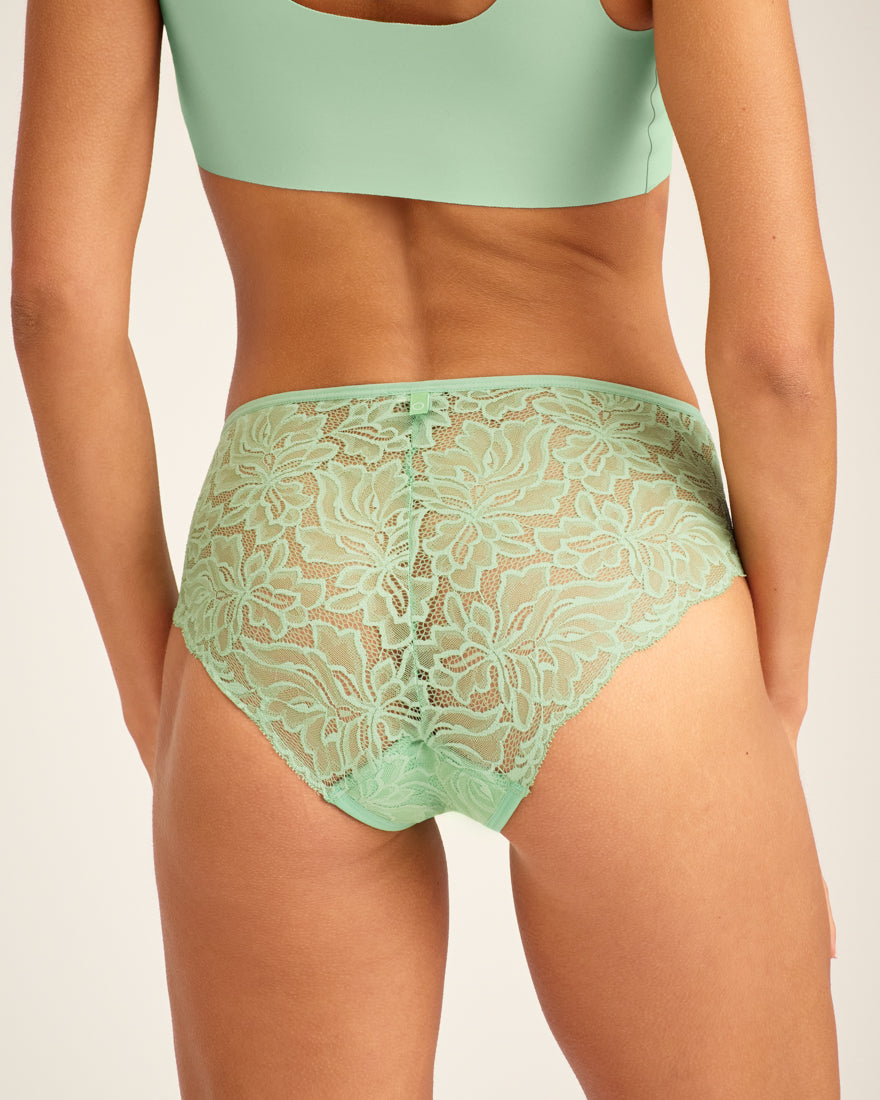 Modell trägt ooia Hipster Allover Lace in Bright Sage – Periodenslip mit durchgehender floraler Spitze. Rückansicht.
