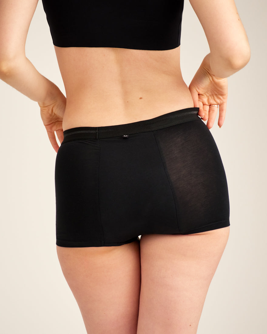 Modell trägt ooia Shorty Extra Strong in Schwarz – Periodenunterwäsche mit breitem Bund und starker Saugstärke. Rückansicht.