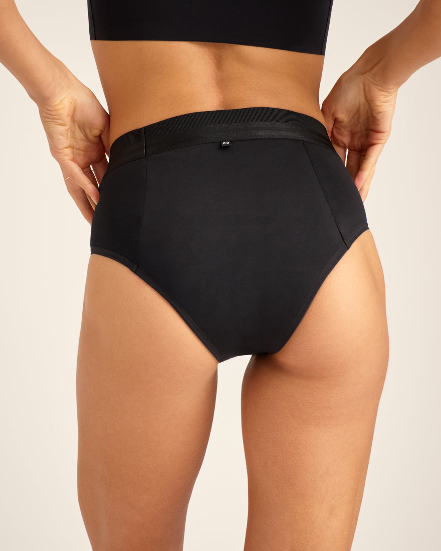 Modell trägt ooia Slip Hugs Extra Strong in Schwarz – bequemer Slip mit extra stützendem Bund, starker Sauglstärke und integrierten Taschen für das ooia Wärmepad. Rückansicht.