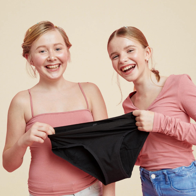 Zwei Mädchen halten lächelnd zusammen einen schwarzen Menstruationsslip für Teens in den Händen