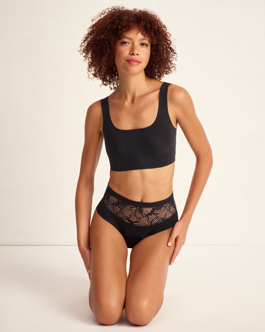 Modell trägt Periodenunterwäsche Highwaist Seamless Lace extra stark in schwarz mit nahtlosem Design mit breiter Spitze am Bund.
Frontal, kniend